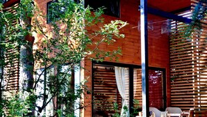 Un edificio moderno de madera con ventanas amplias, un espacio de estar cubierto al aire libre y un árbol en primer plano.