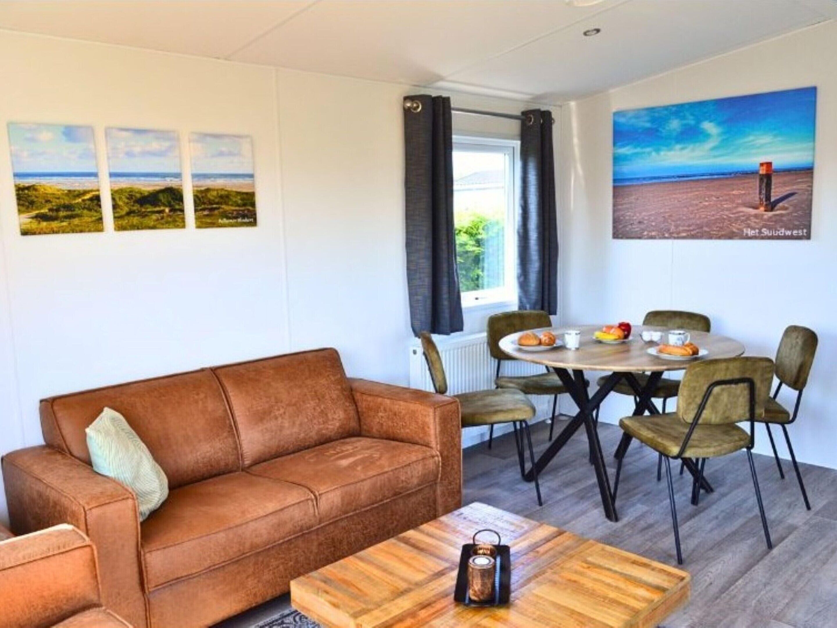 Une pièce chaleureuse avec un canapé en cuir marron, une table basse en bois et une table à manger avec des chaises. Trois tableaux encadrés ornent le mur, il y a une fenêtre avec des rideaux et un tableau représentant une scène de plage.