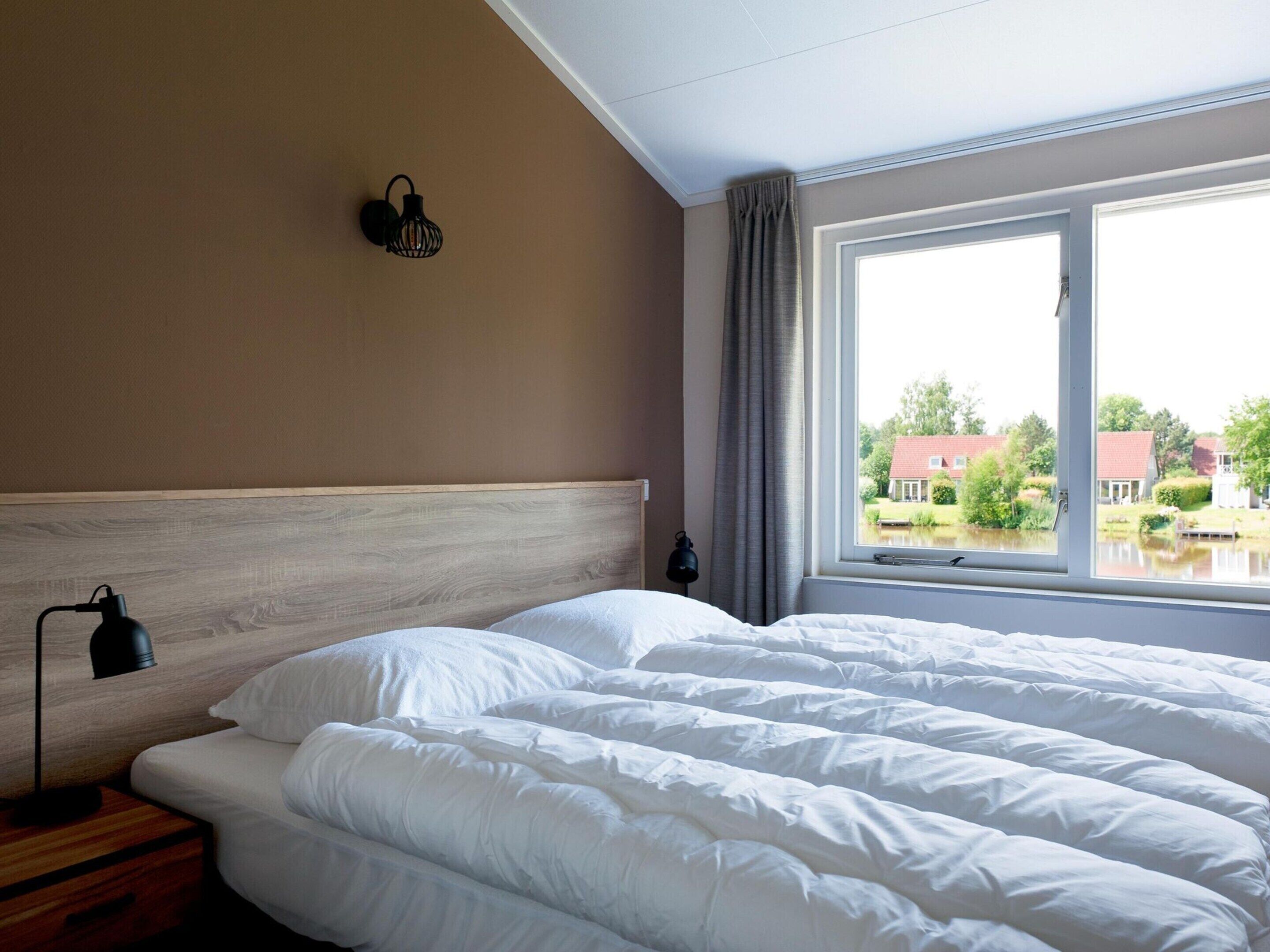 A hotel room with a large bed, a wooden headboard, a nightstand, a lamp, and a window with a view of houses and greenery.
