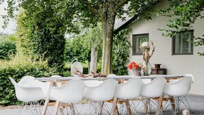 An outdoor dining area with a long table and white chairs, surrounded by greenery and a white brick building. - House in Leenderstrijp Near Leenderbos Trails (Leende)