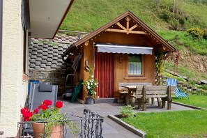 A wooden cabin with a stone wall entrance, a wooden bench, a table, and a chair outside. - Salzburg Lungau Nature Apartment (Muhr)