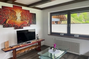 Uma sala de estar com televisão, um banco de madeira, mesa de centro de vidro e obras de arte na parede.
