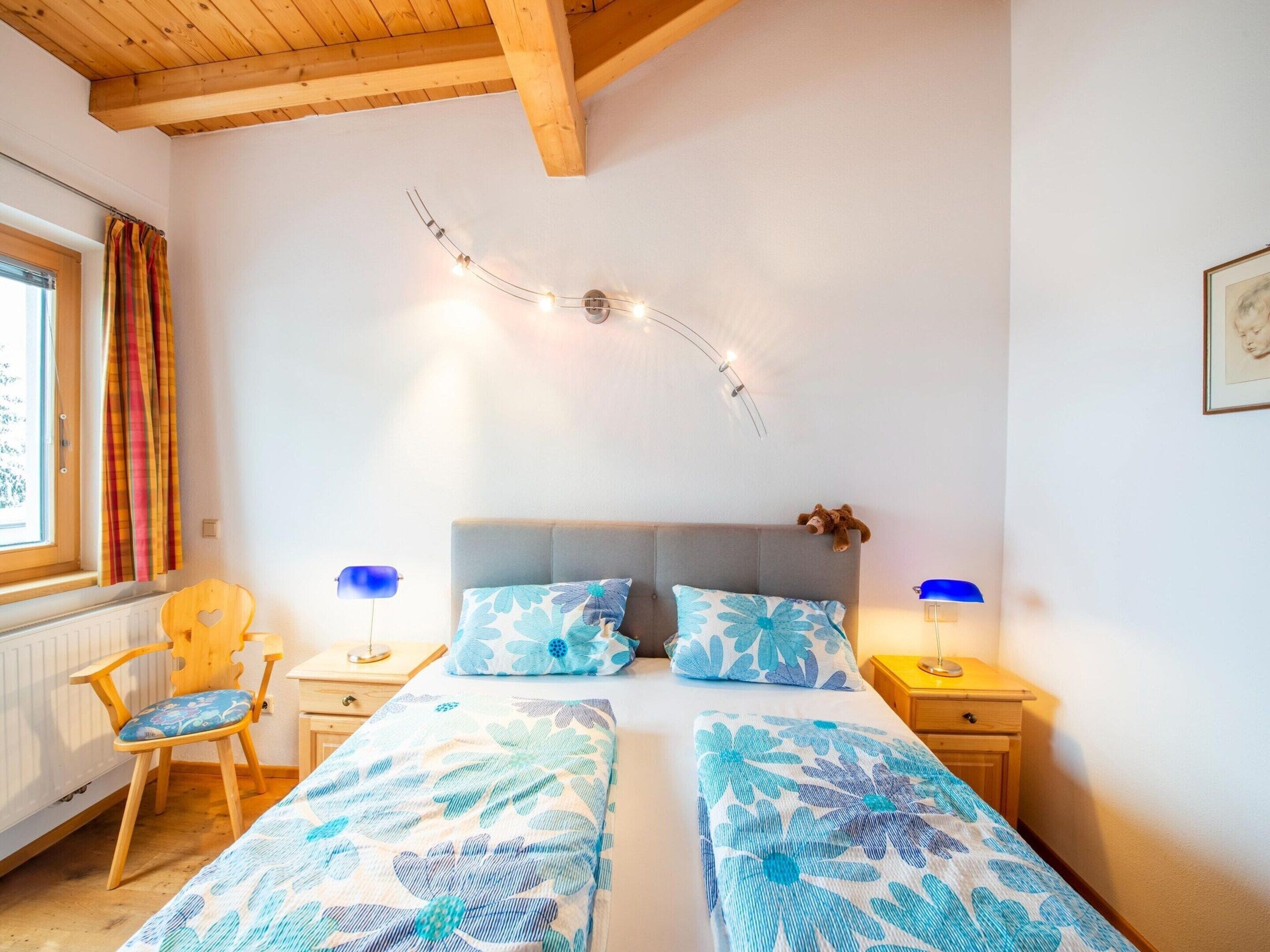 A bedroom with two beds, a wooden chair, a nightstand with a lamp, and a window with curtains.