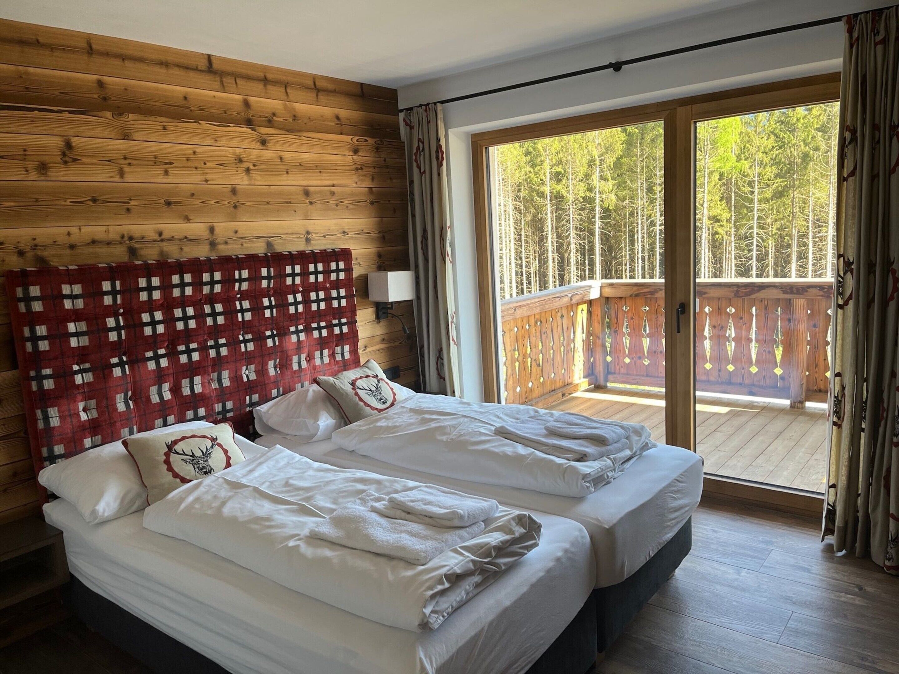A room with two beds, wooden walls, a balcony with a view of trees, and a red and white checkered headboard.