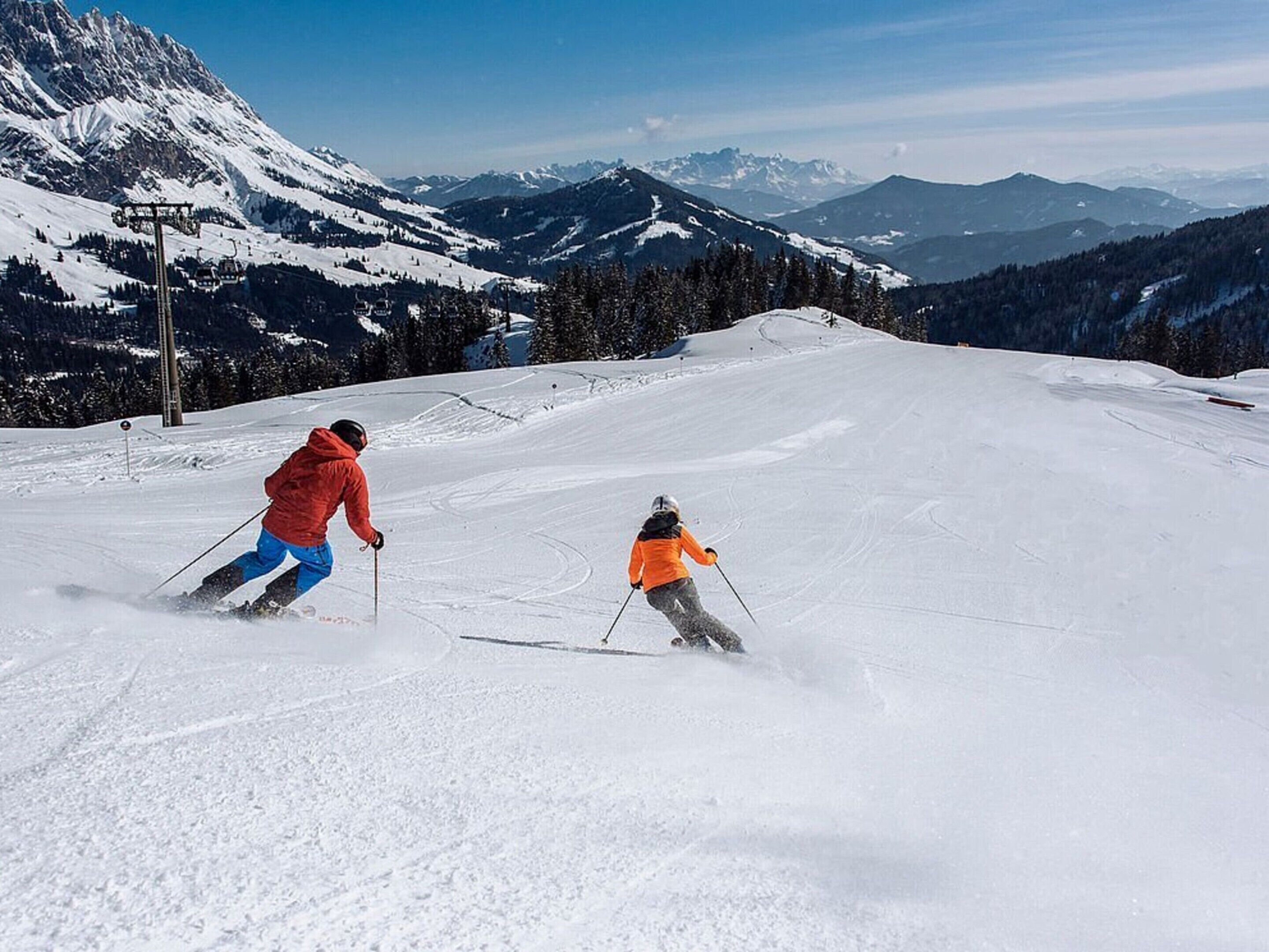 A ski slope with two skiers, one in red and one in orange, with snow-covered mountains in the background.