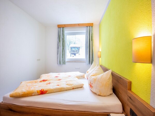 A single bed with floral bedding, a window with curtains, and a wall-mounted lamp.