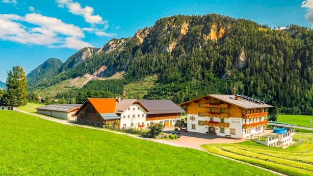 Un edificio de estilo alpino tradicional con un amplio balcón, rodeado de campos verdes exuberantes y montañas al fondo.