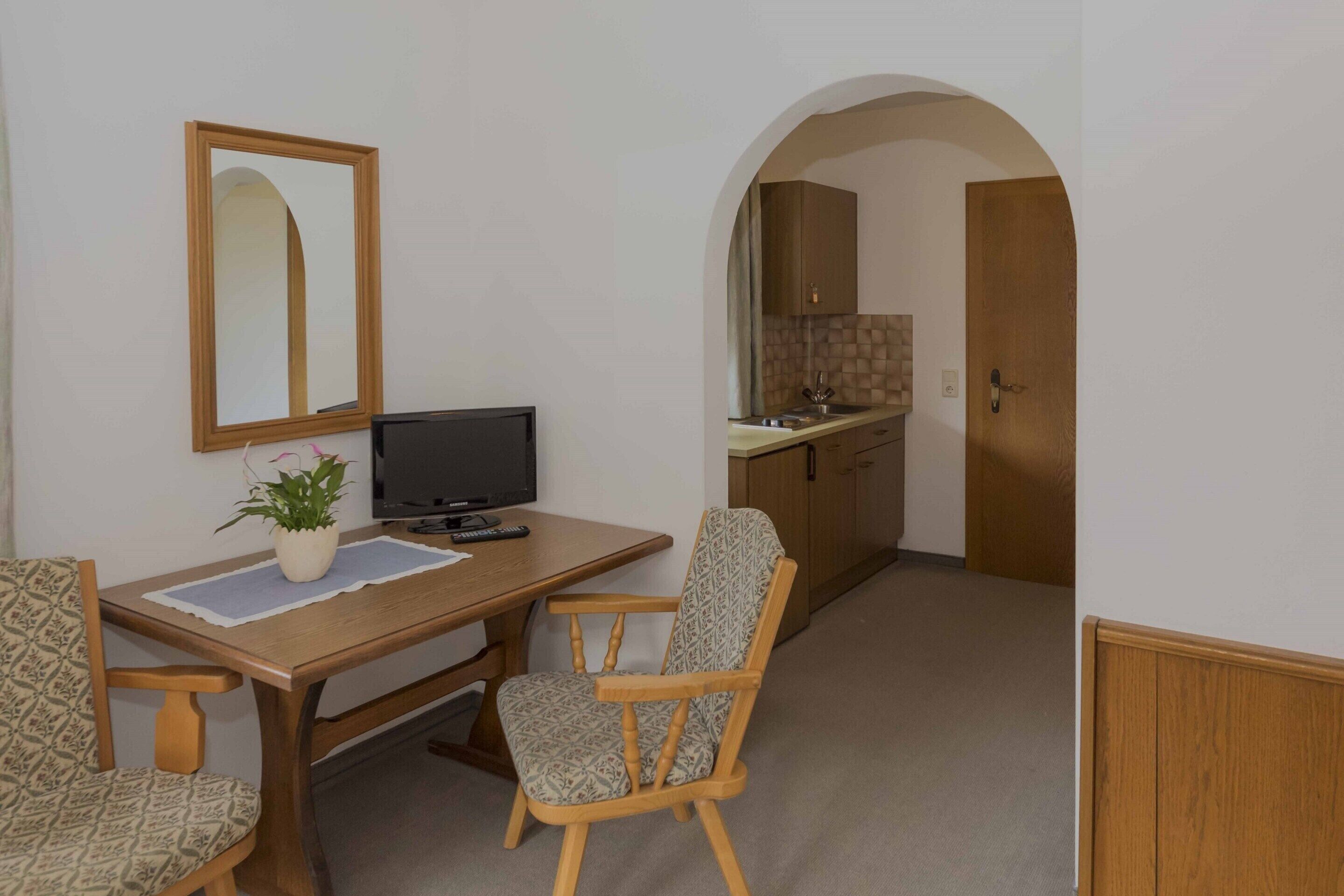 A small room with a wooden desk, a television, a chair, a potted plant, and a mirror. There is a doorway leading to a kitchen area.