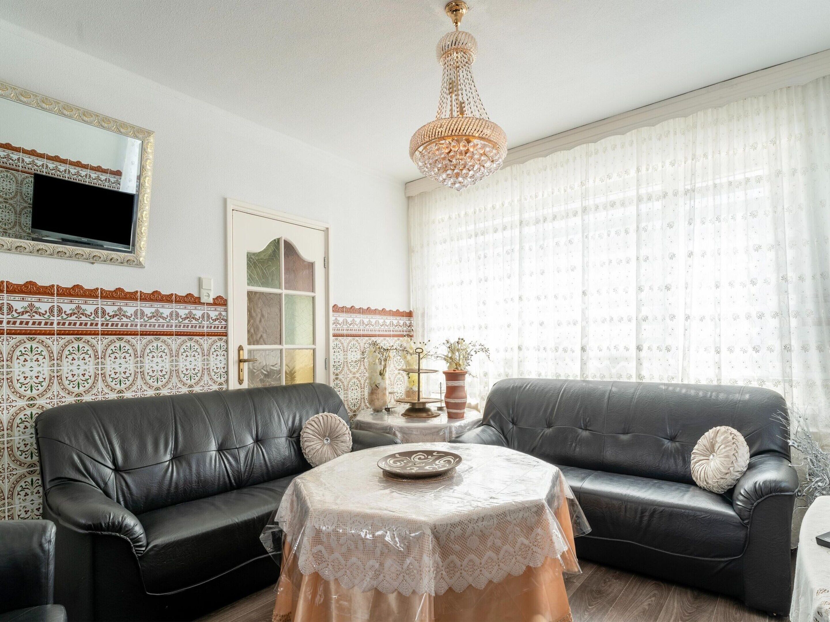 Ein Wohnzimmer mit zwei schwarzen Ledersofas, einem durchbrochenen Couchtisch, einem dekorativen Kronleuchter, Wandfliesen mit Muster und einem großen Fenster mit durchsichtigen Vorhängen.