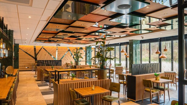 A modern restaurant interior with wooden furniture, a central plant, and large windows offering views.