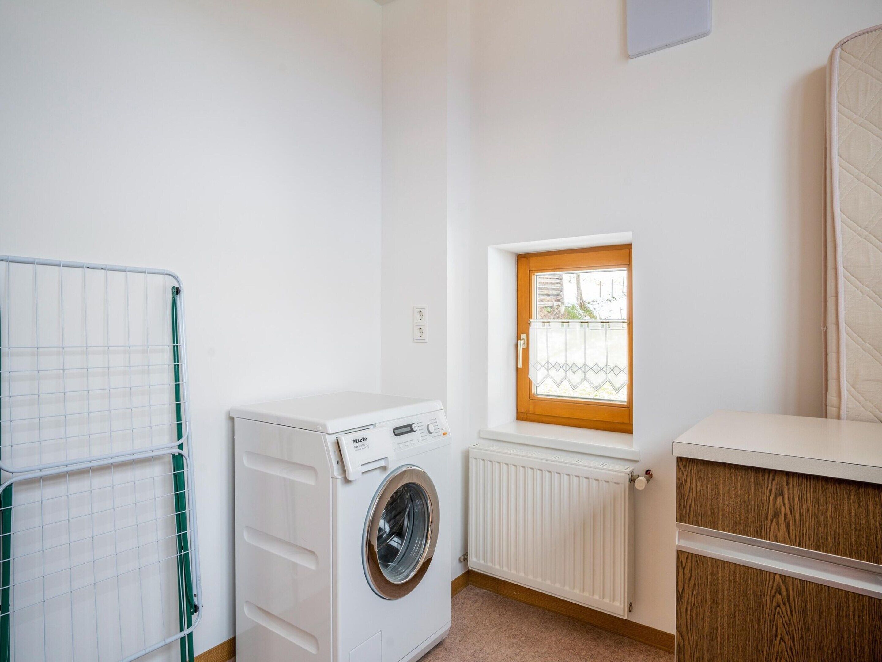 A laundry room with a front-loading washing machine, a drying rack, a window with a view, and a radiator.