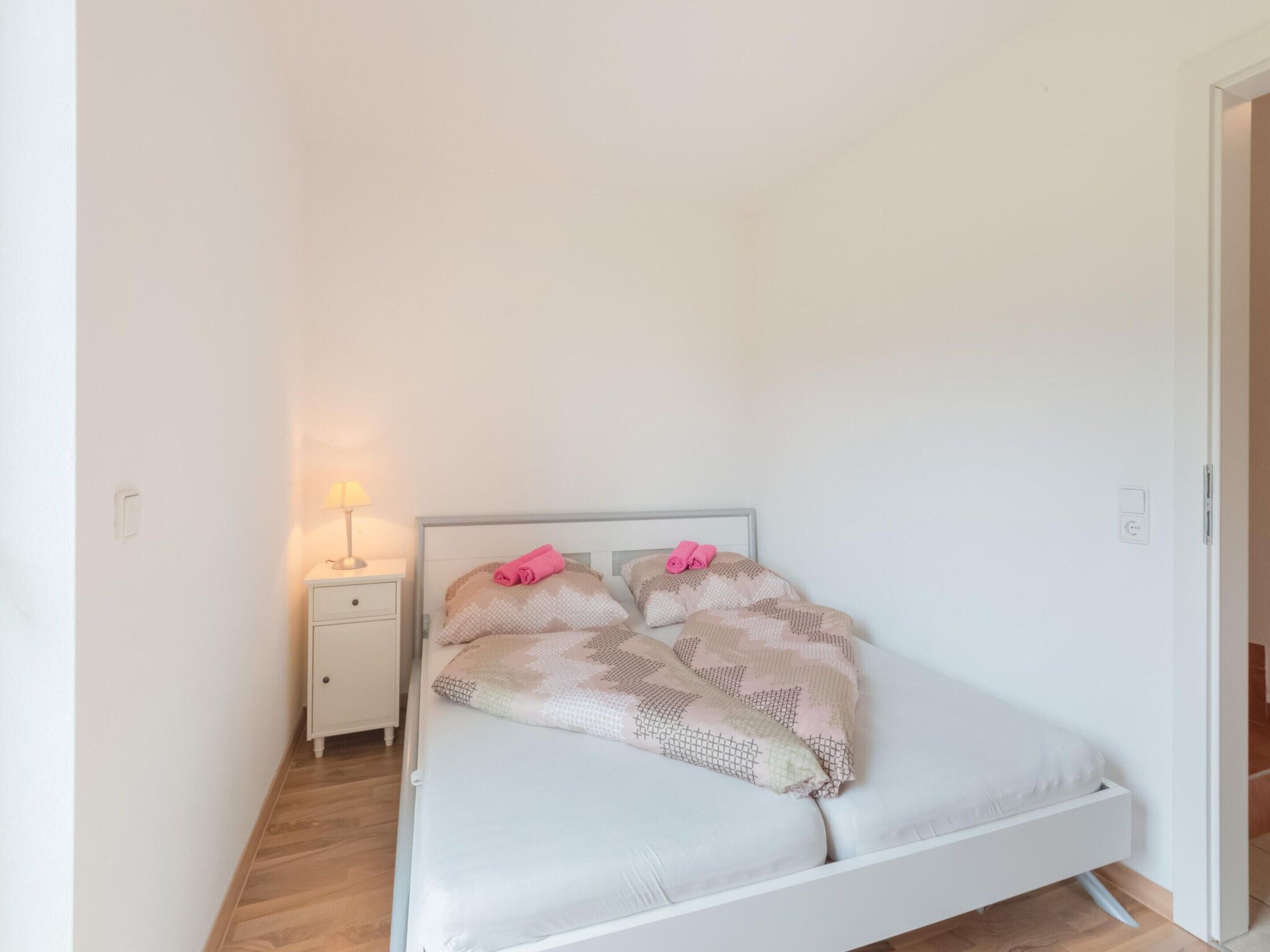 A small, single-bed room with a white bed, a nightstand with a lamp, and a plain white wall.