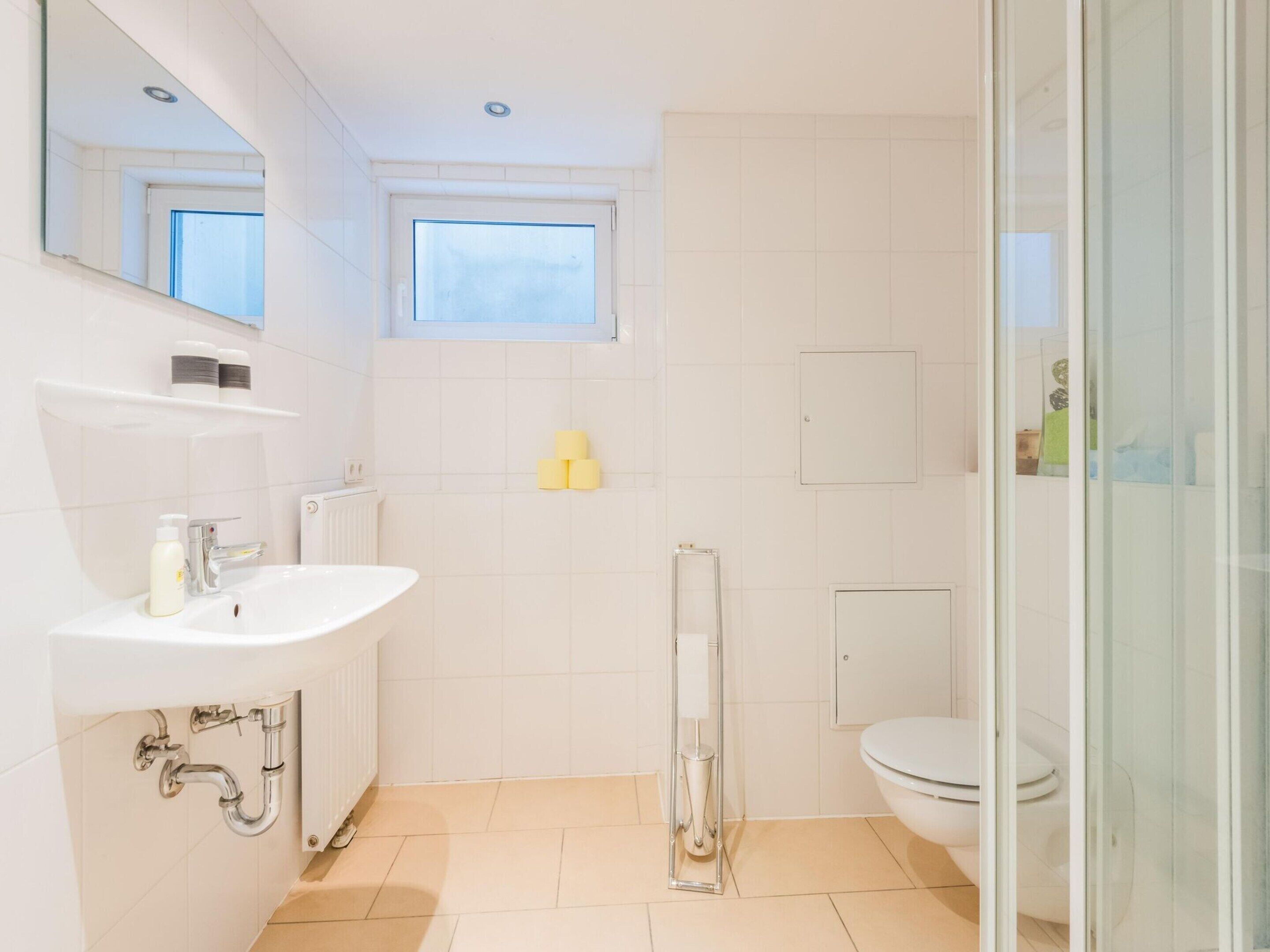 Un bagno moderno con lavandino, specchio e servizi igienici bianchi. Sopra il lavandino si trova una finestra e sulla destra è presente la doccia.