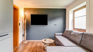 A modern living room with a grey sofa, a flat-screen TV mounted on the wall, a wooden floor, and a small round table with a plant. - High-quality Apartment With Whirlpool (Bramberg am Wildkogel)
