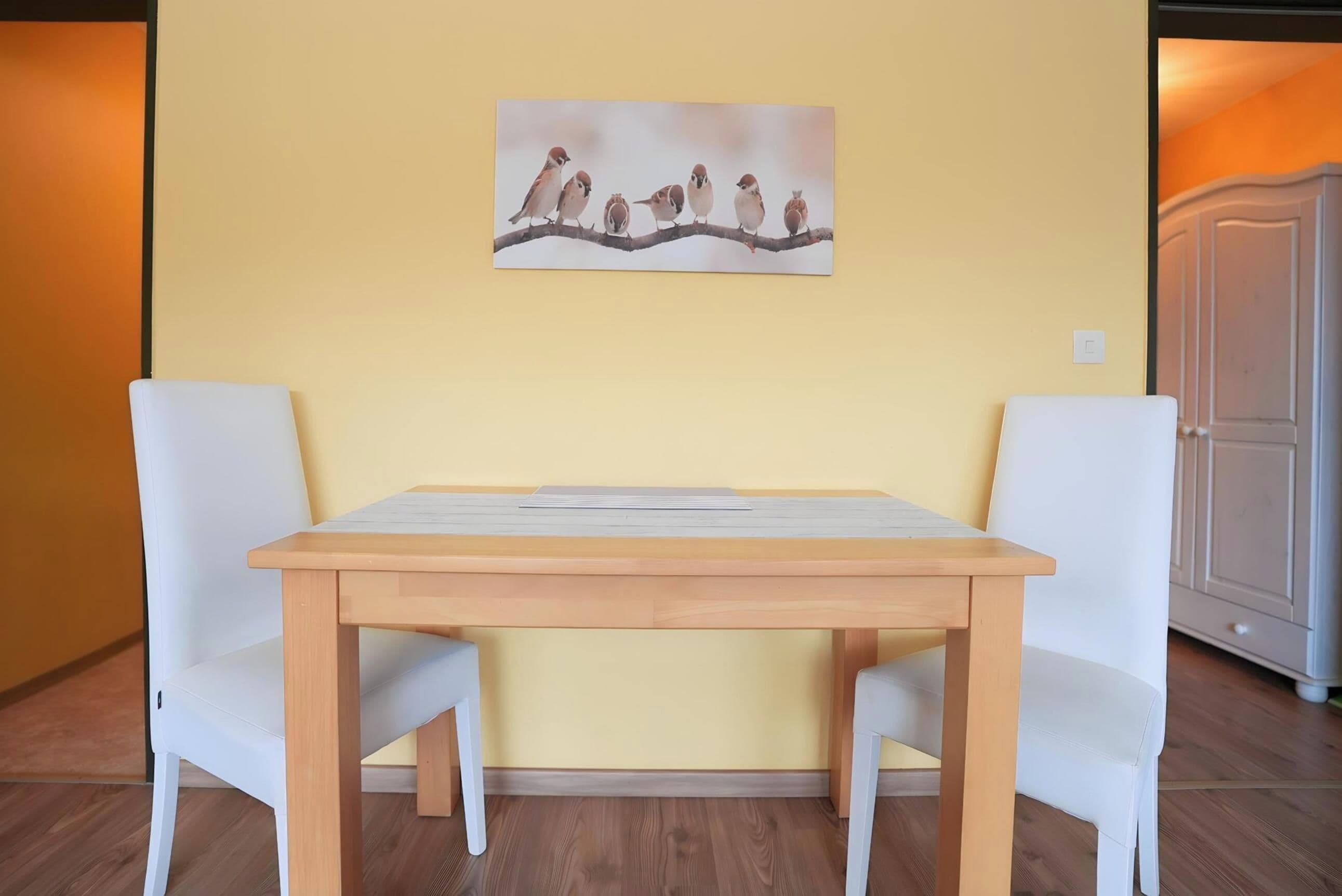A dining area with a wooden table and two white chairs, a wall-mounted artwork above the table, and a door to the right.