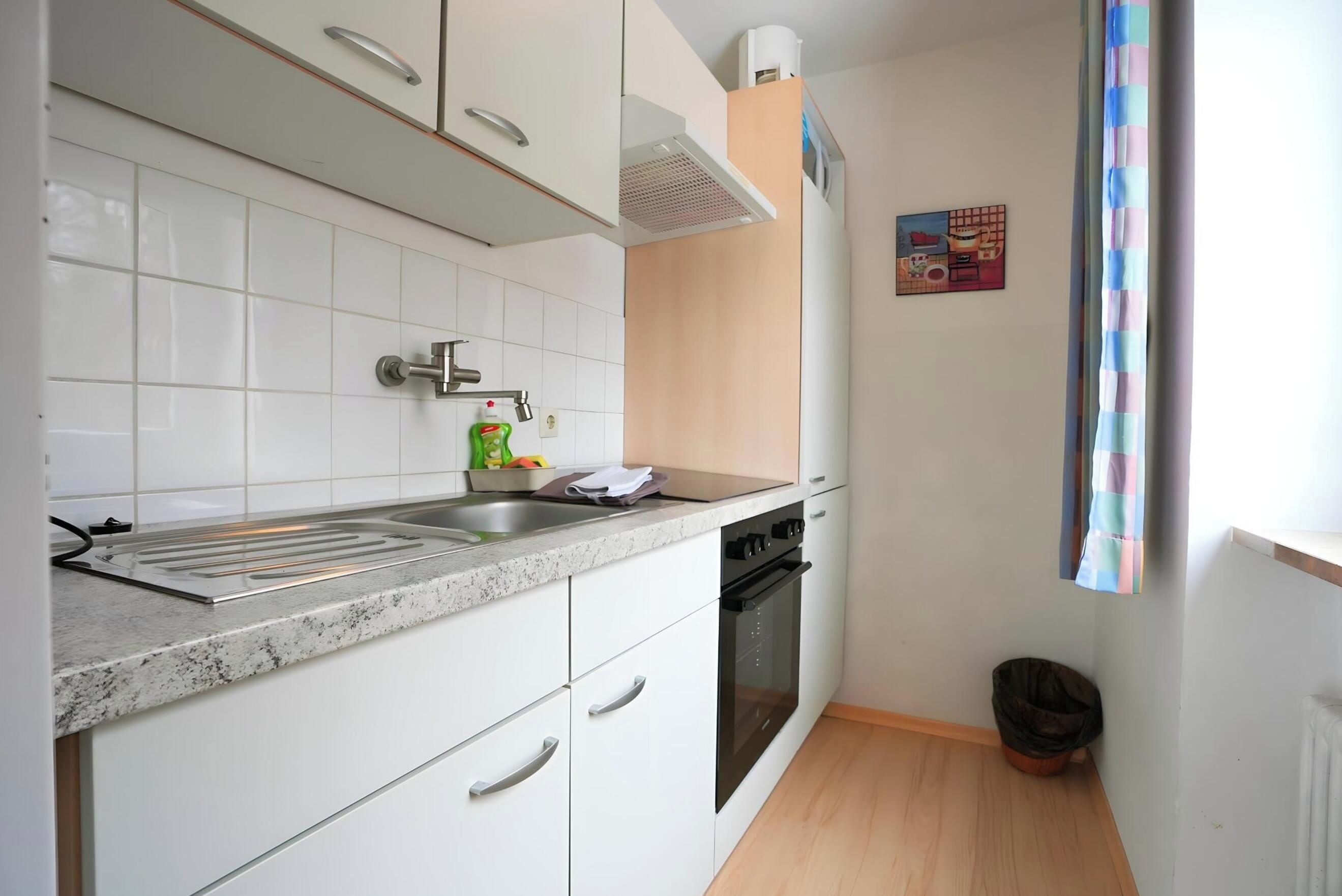 A kitchen with white cabinets, a stainless steel sink, a built-in oven, and a window with a striped curtain.