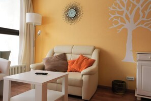 Un salon comprenant un canapé, une table, une lampe, une horloge murale et un motif décoratif représentant un arbre.