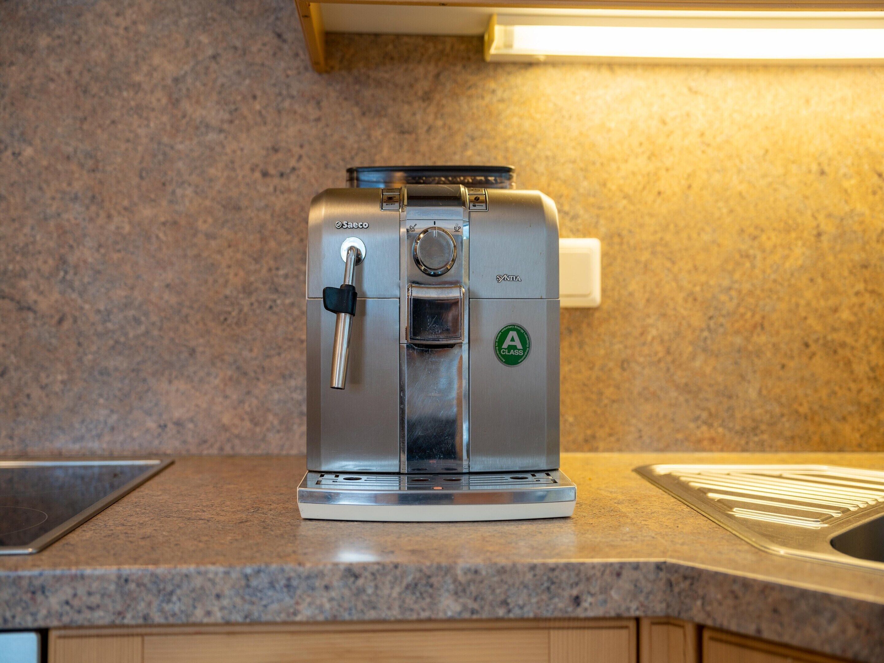A stainless steel espresso machine on a granite countertop with a sink and cabinets in the background.