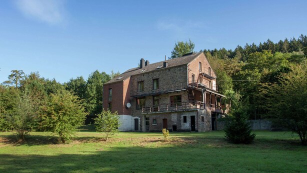 Een groot, stenen gebouw met twee verdiepingen en een balkon op de bovenverdieping, omringd door bomen en een gazon.
