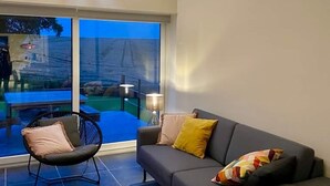 Ett modernt vardagsrum med en grå soffa, en svart fåtölj och ett stort fönster som ger utsikt över en uteplats med ett bord och stolar.