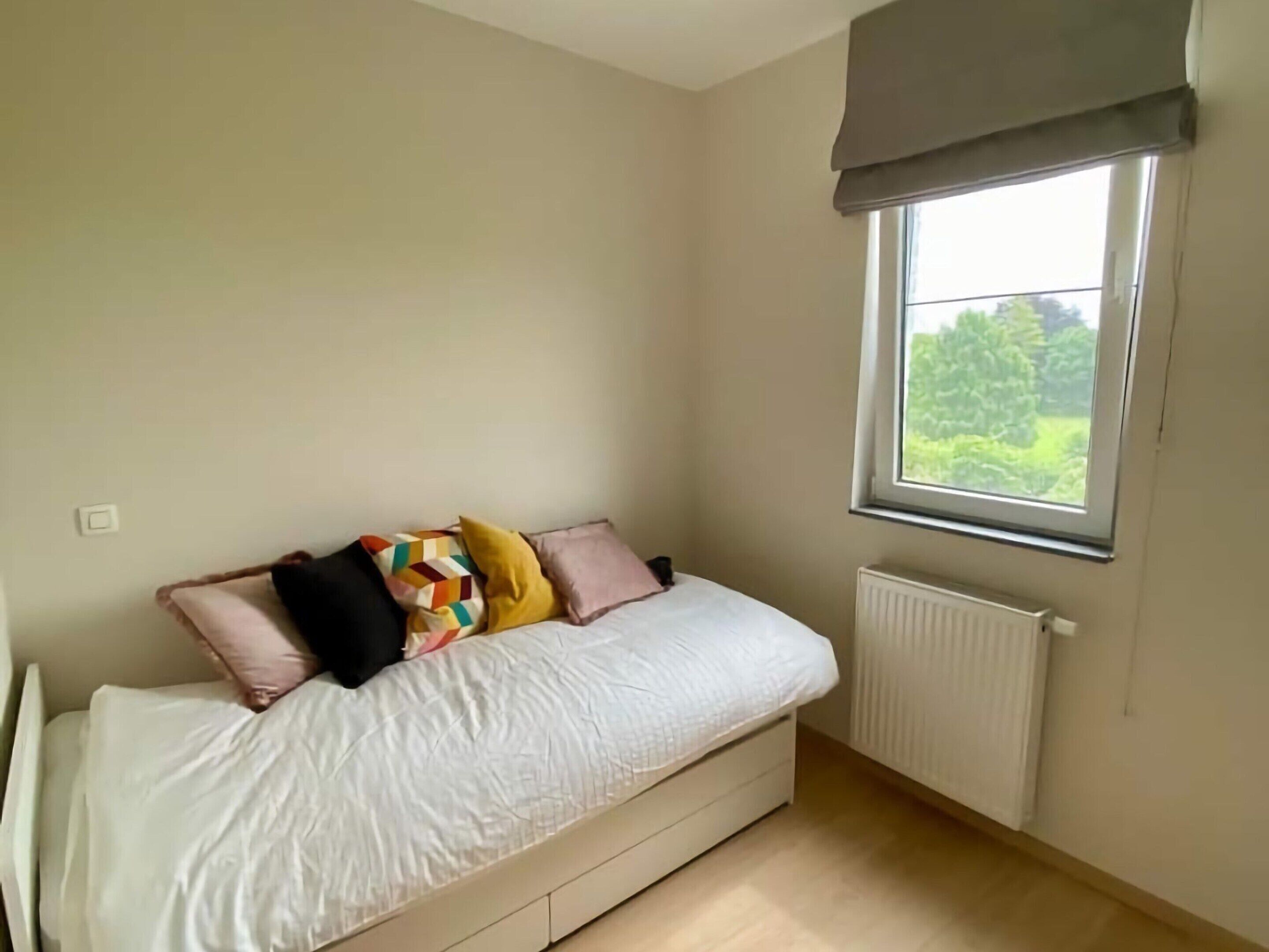 A bedroom with a bed, a window with a view of greenery, and a radiator.