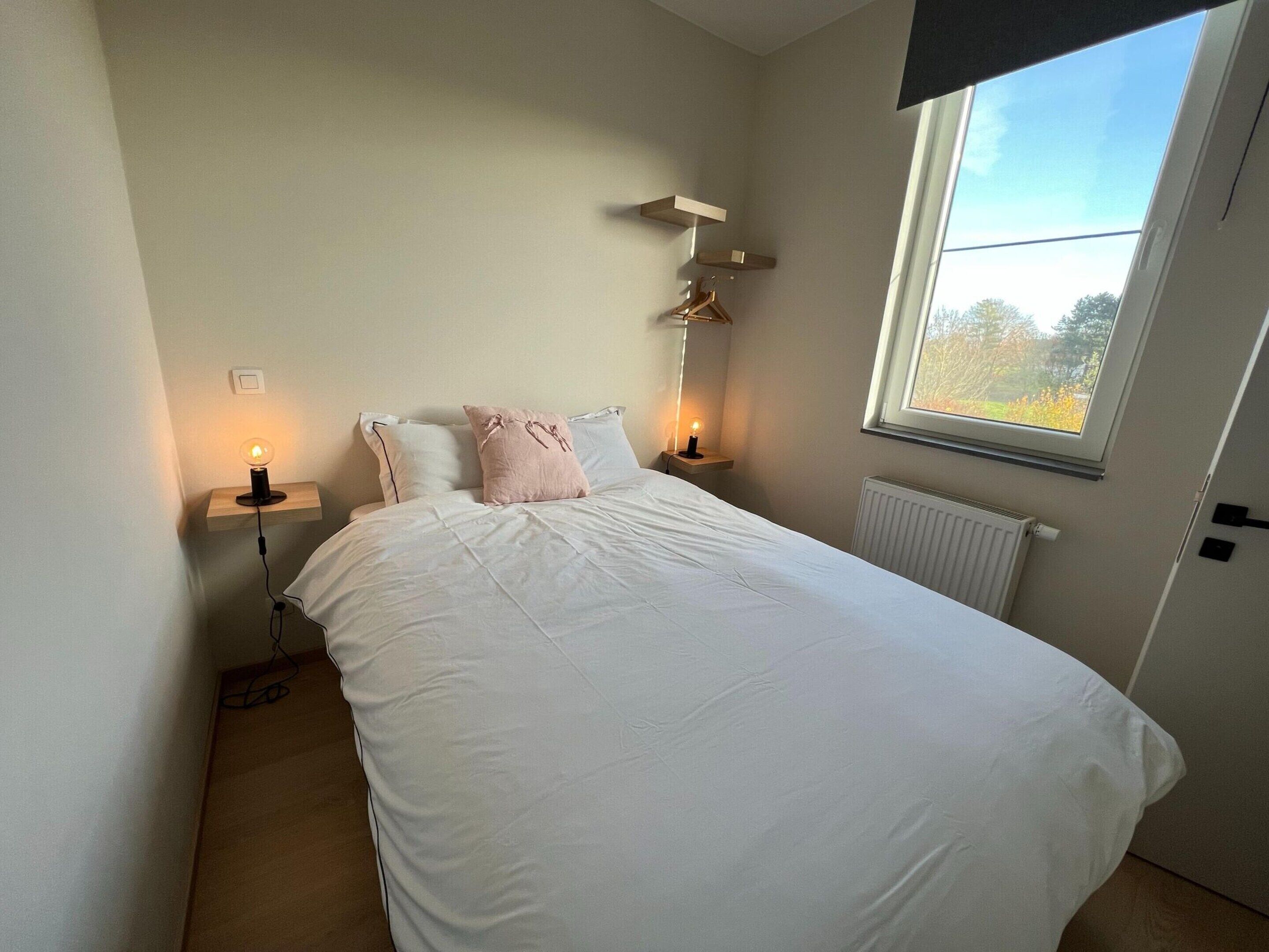 A neatly made bed with white linens and two pillows, a bedside table with a lamp, and a window with a view of trees.