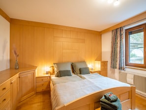 A bedroom with a wooden headboard, two bedside tables with lamps, a bed with a white duvet, a radiator, and a window with curtains. - Ferienwohnung mit Fun+ Card in Niedernsill (Niedernsill)