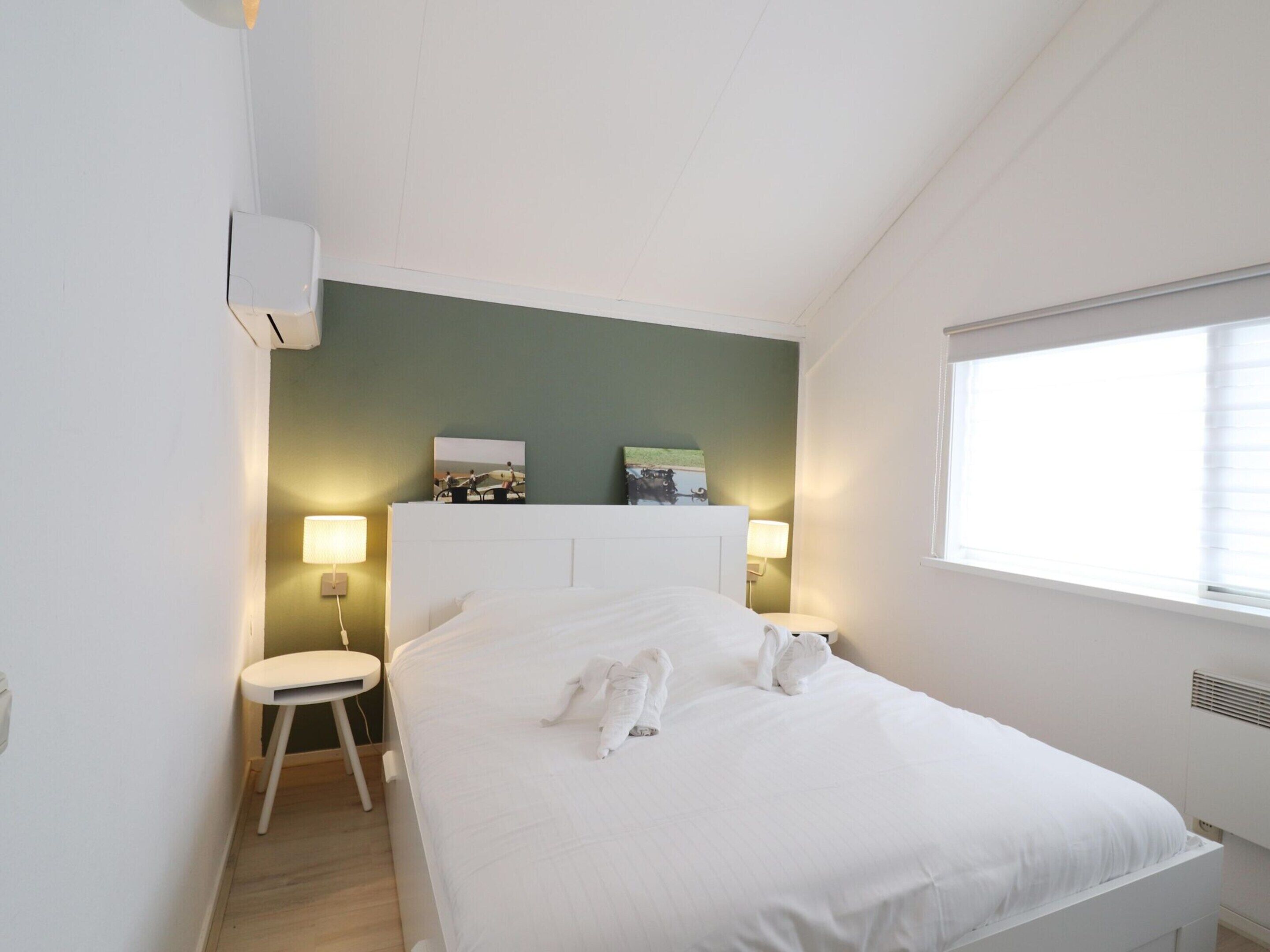 A bedroom with a bed, two bedside lamps, a small stool, and a wall-mounted air conditioner.