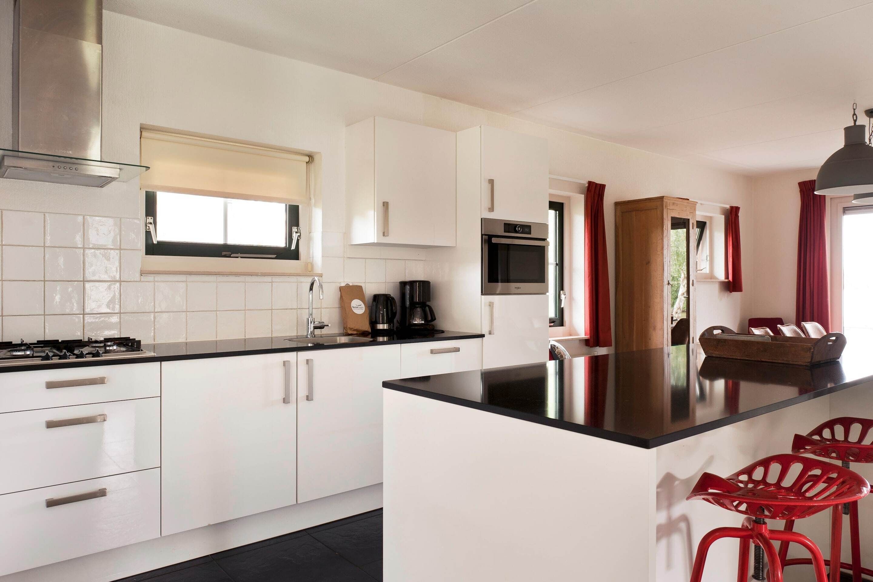 A modern kitchen with white cabinets, a black countertop, a stainless steel range hood, a built-in oven, and a coffee maker. There are red bar stools and a wooden cabinet with a mirror.