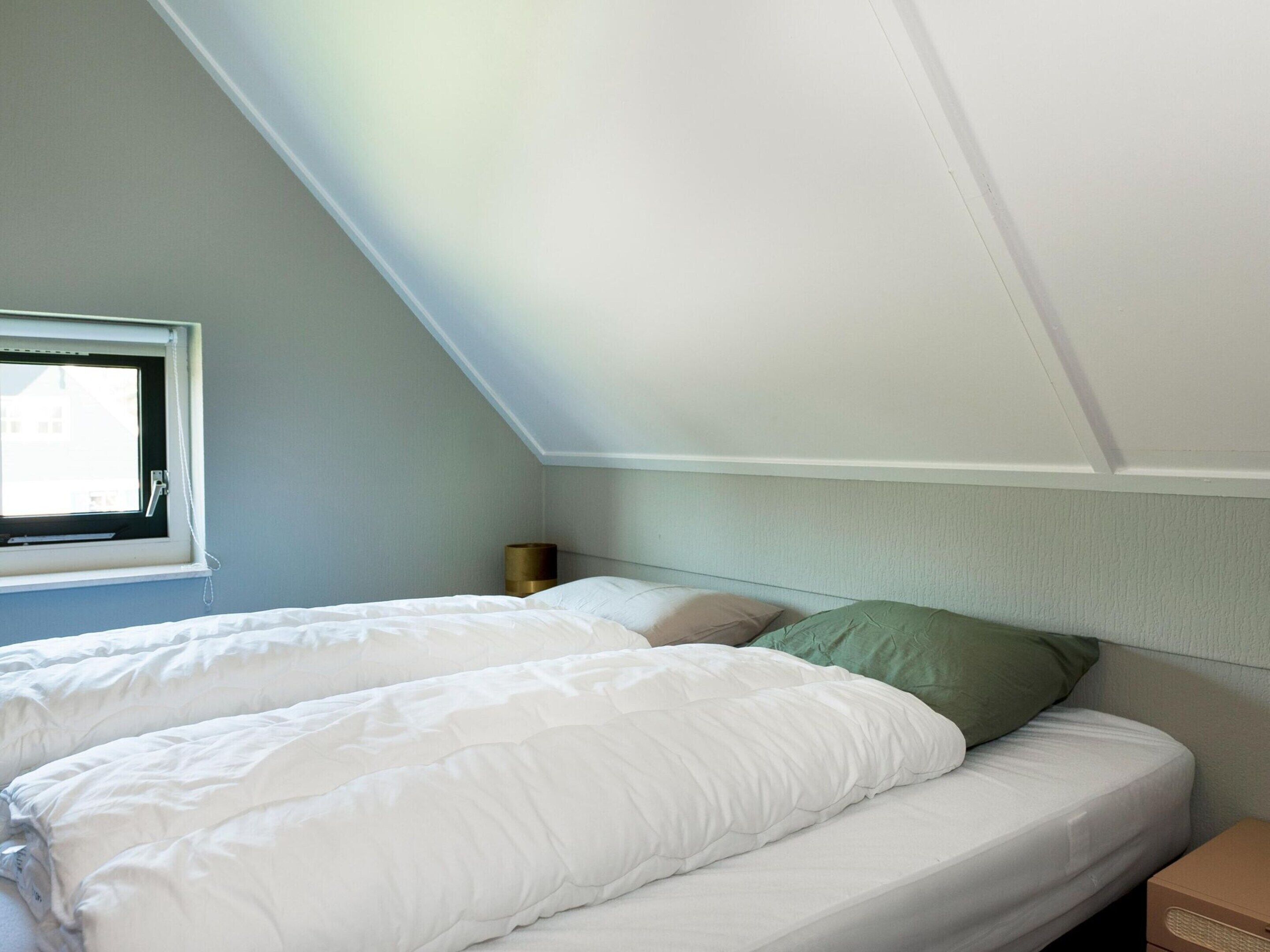 A bedroom with a sloped ceiling, a window, a bed with white bedding, a green pillow, and a bedside table.