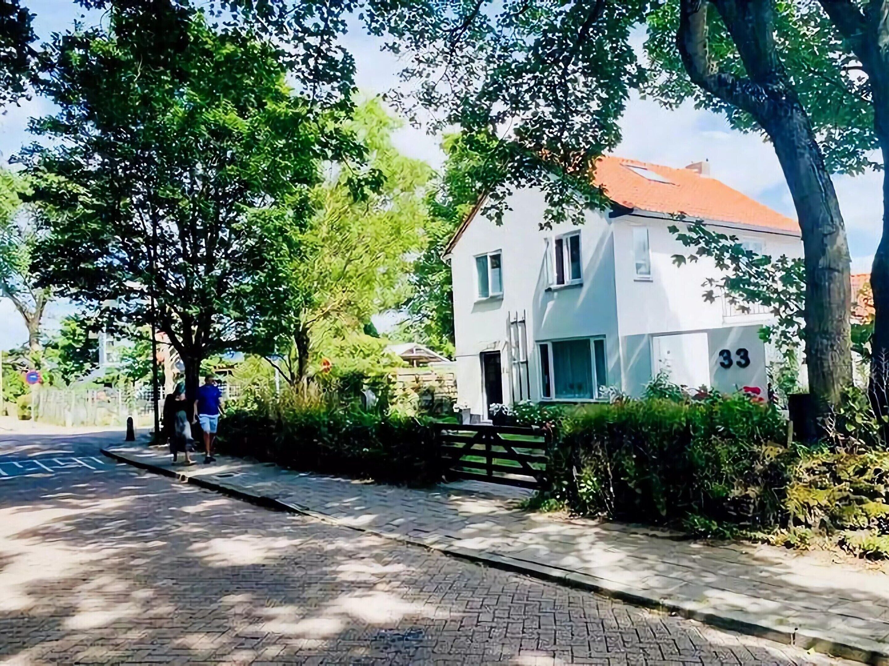 A residential street with a white house numbered 33, paved sidewalks, and lush greenery.
