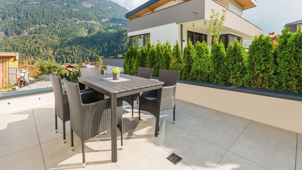 Ein modernes Haus mit Terrasse, ausgestattet mit Esstisch und Stühlen, umgeben von Grün und mit Blick auf eine Berglandschaft.