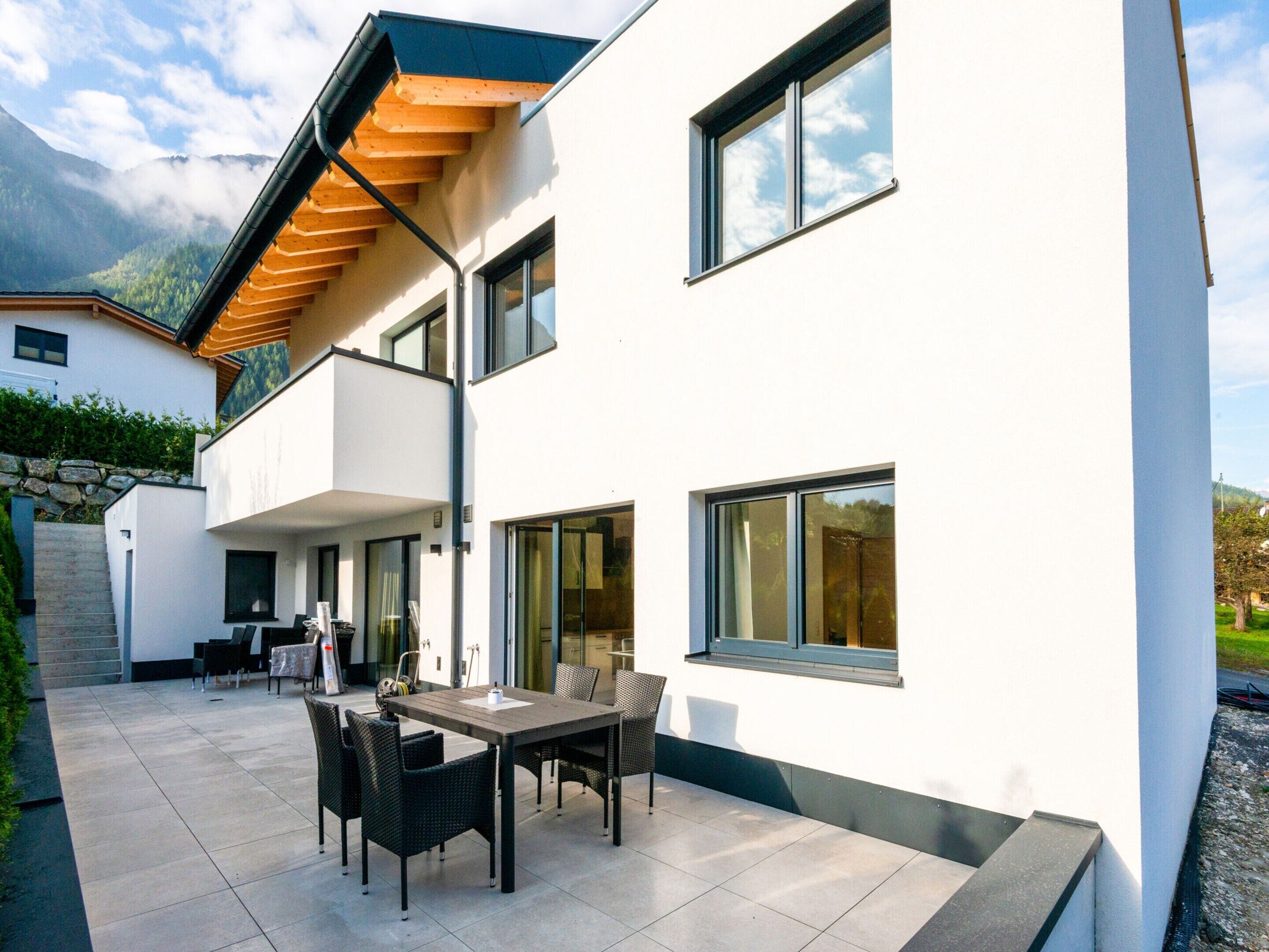 Ein modernes, zweistöckiges Haus mit Balkon, großen Fenstern und einer Terrasse mit Gartenmöbeln.