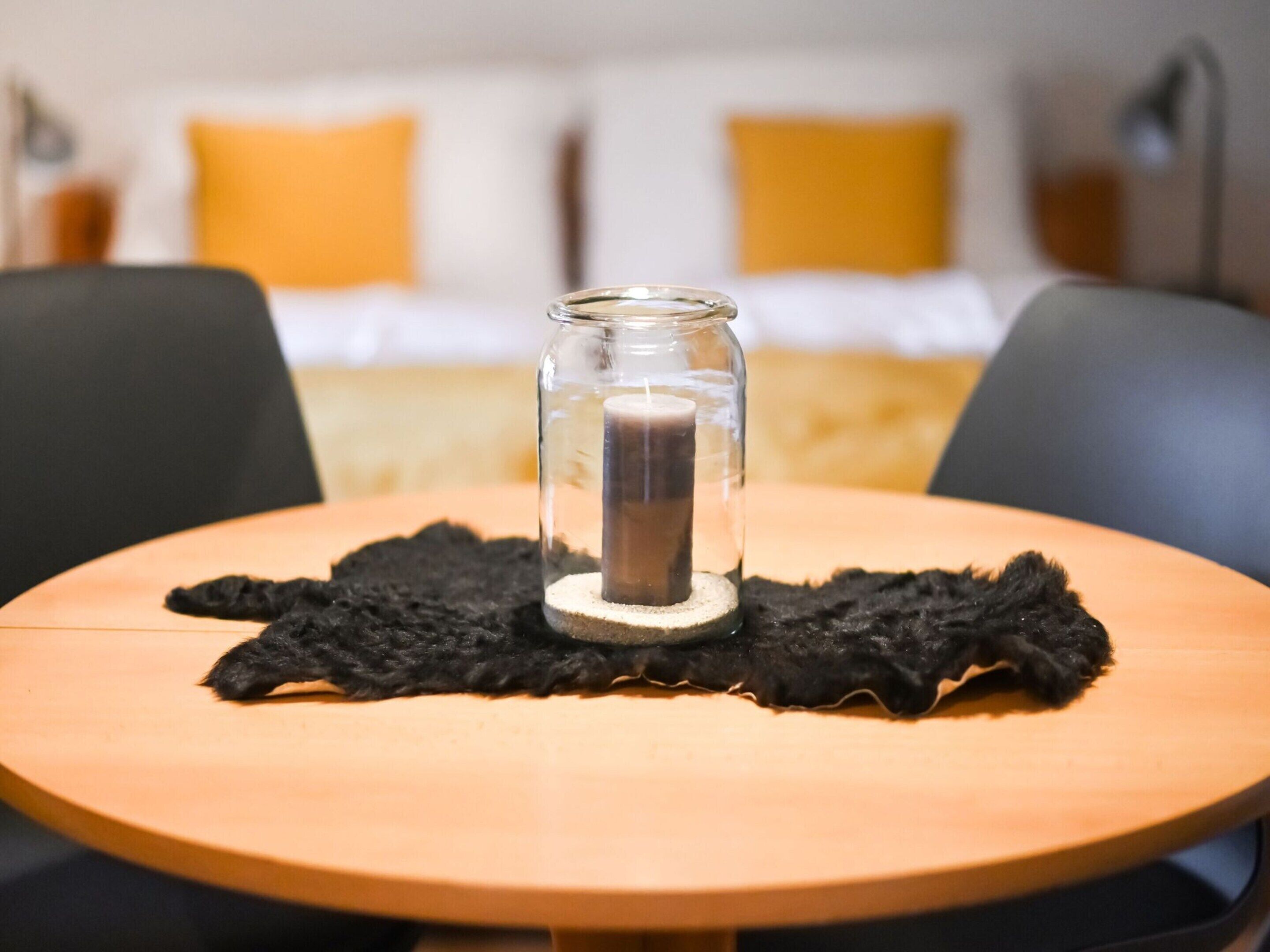 Una mesa redonda de madera con un candelabro de vidrio que contiene una vela, un posavasos negro con textura y dos sillas negras.