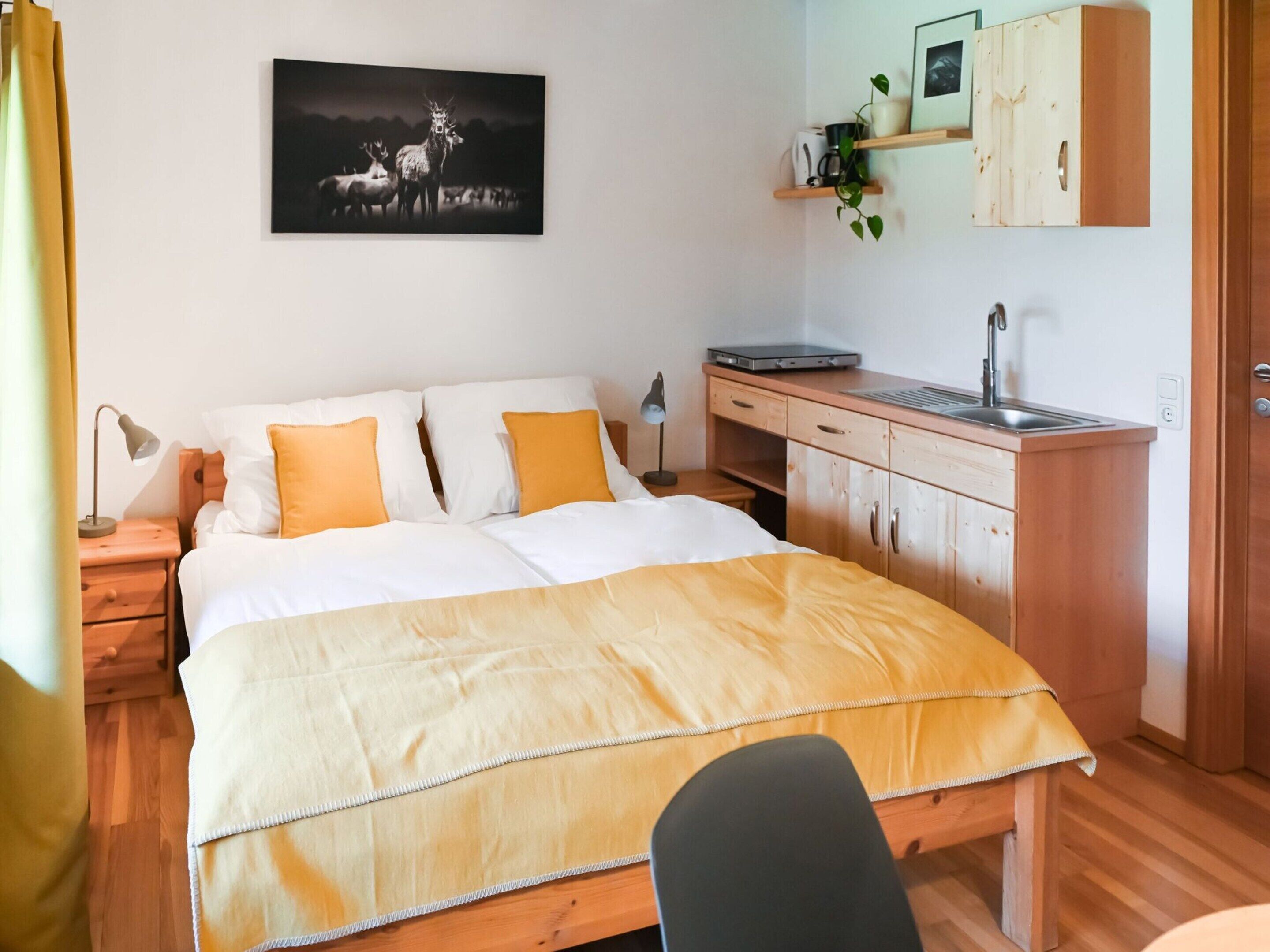 A bedroom with a bed, bedside table, lamp, desk, and a small kitchenette.