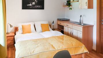 A bedroom with a bed, bedside table, lamp, desk, and a small kitchenette.