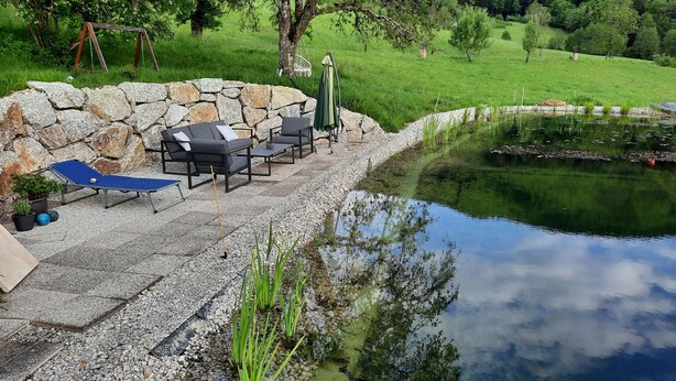 A stone wall with outdoor seating, a pond, and a grassy area with trees. - Loft-like Apartment With Swimming Pond (Scheibbs)
