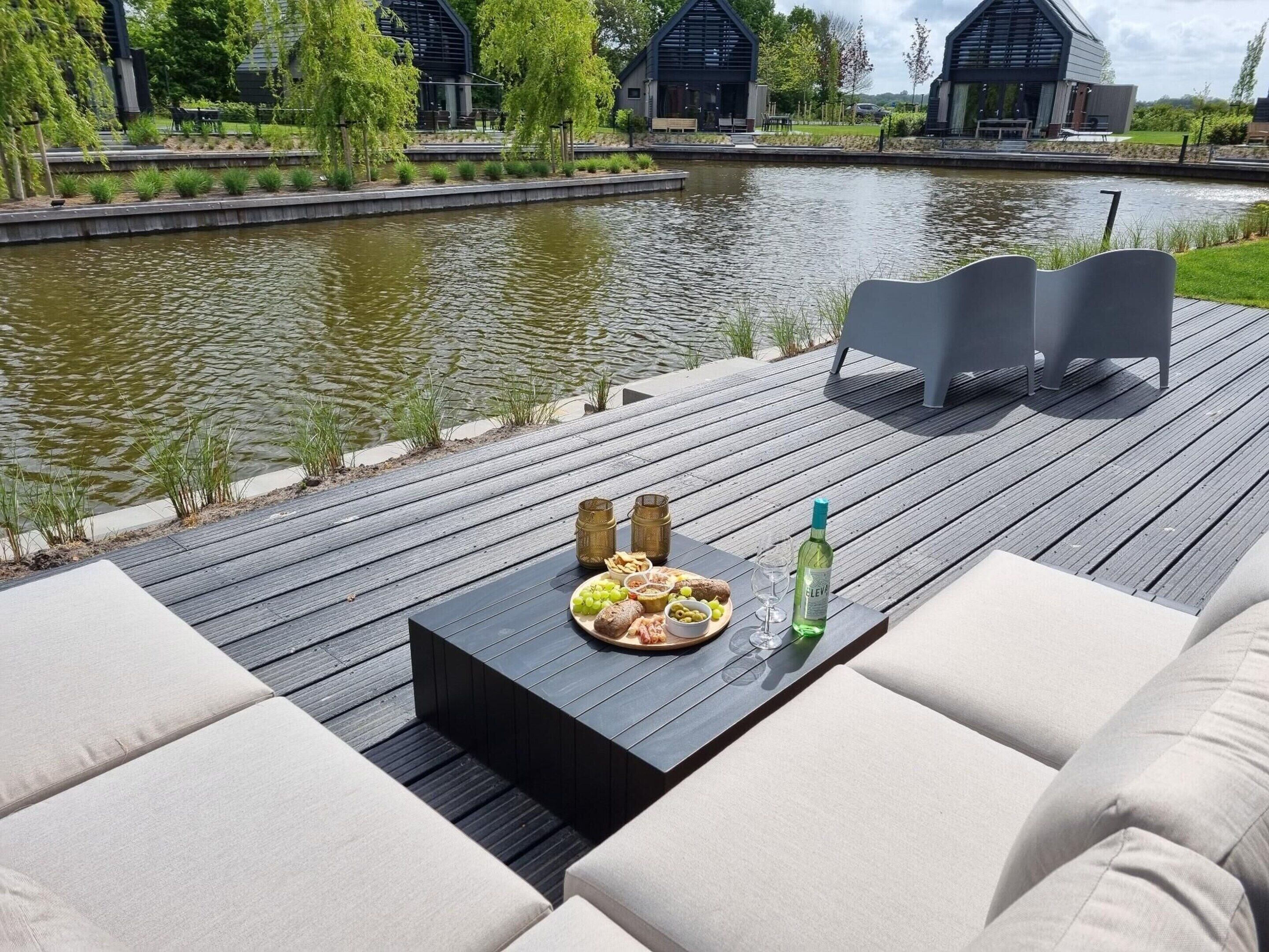 Une terrasse en bois avec une table dressée pour un repas, donnant sur une étendue d’eau calme et sur des bâtiments modernes en arrière-plan.