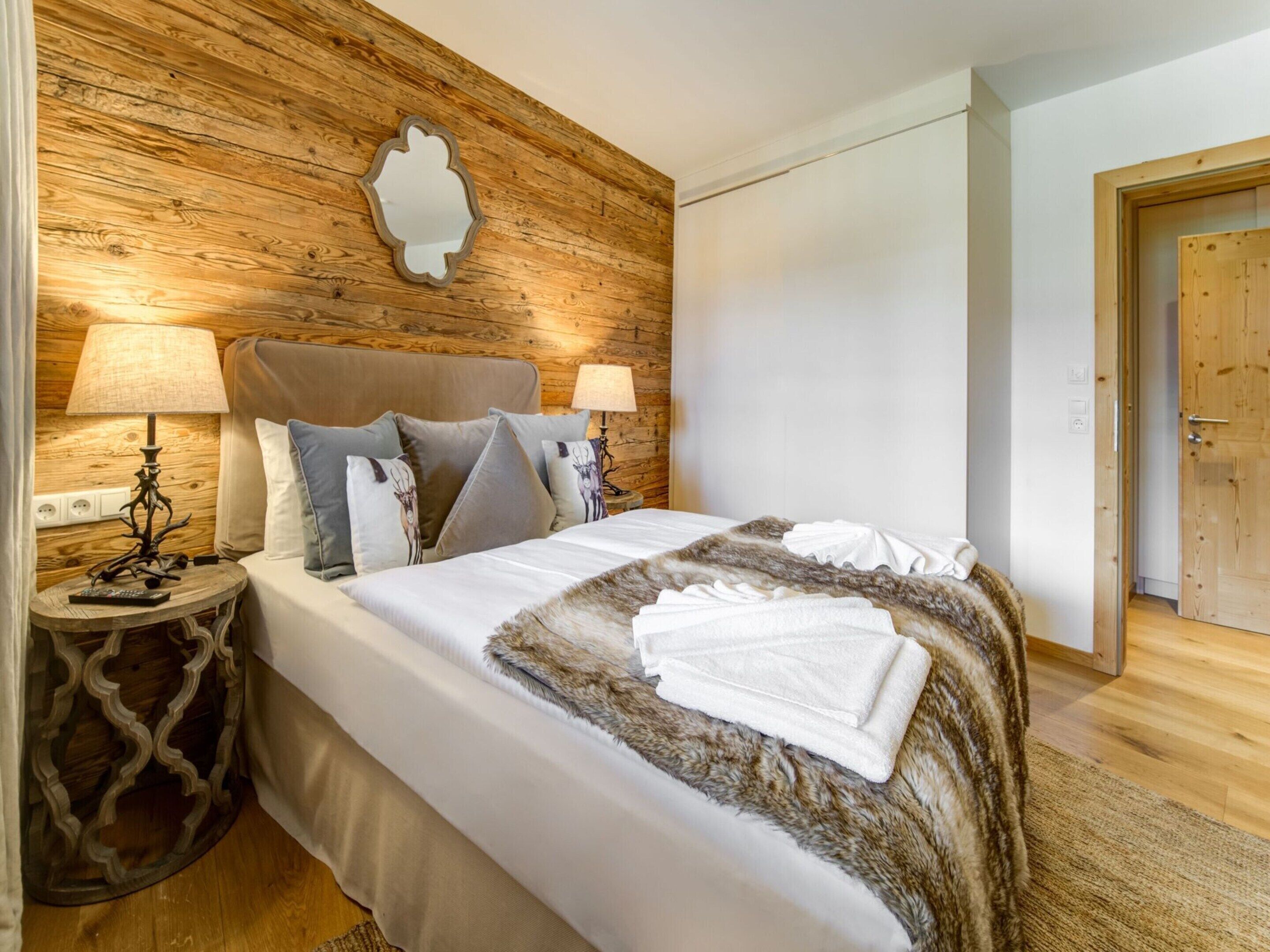 A neatly made bed with a white comforter and a fur throw, flanked by two bedside tables with lamps. The room features a wooden wall panel and a plain white door.