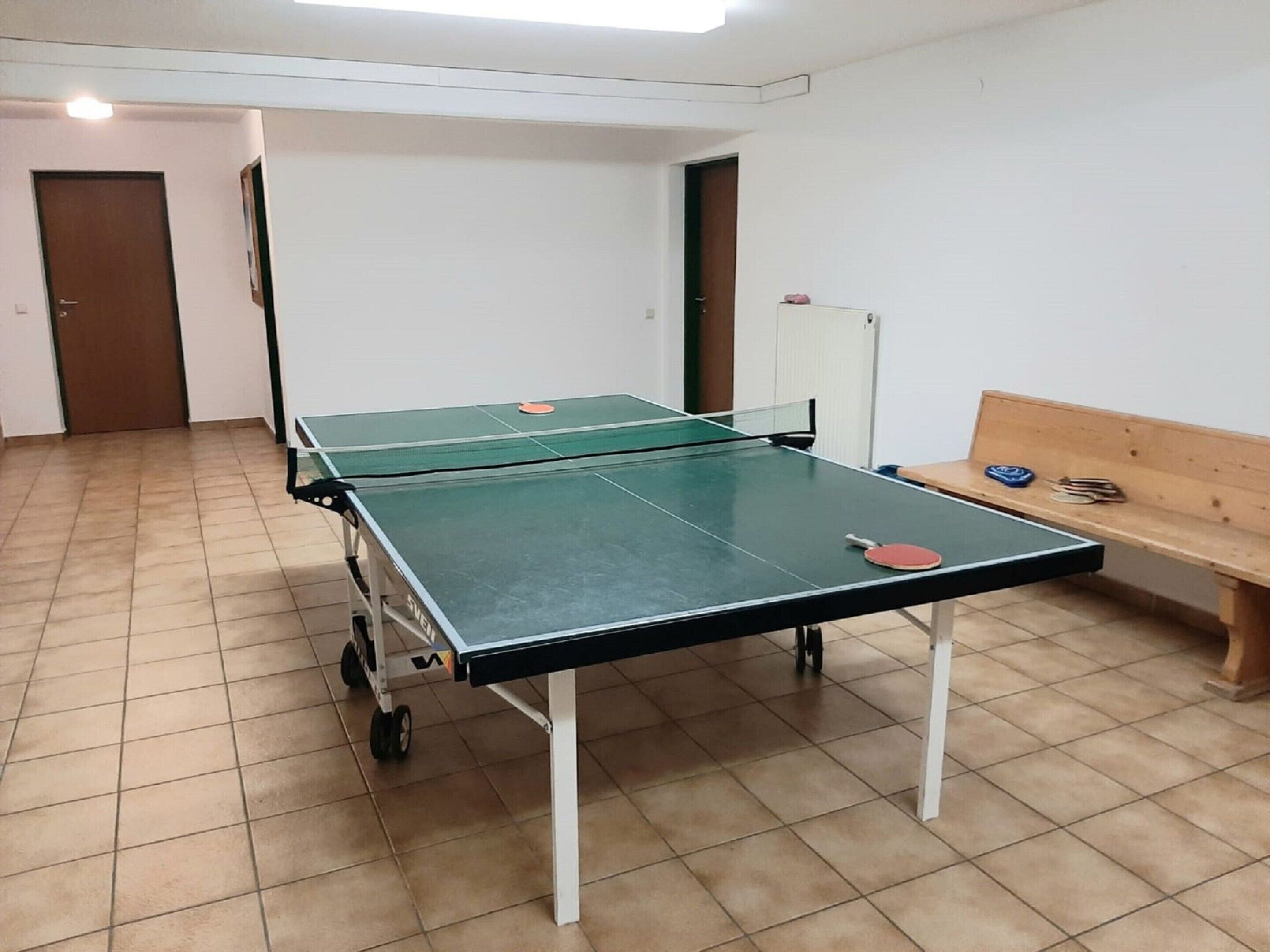Een tafeltennistafel in een ruimte met betegelde vloer, een houten bankje en een deur op de achtergrond.