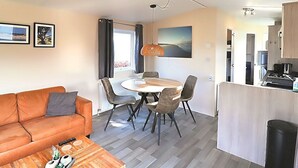 Ein kompakter Wohnbereich mit einer Couch, einem Esstisch, Stühlen, einer Küchenzeile und Blick ins Freie.