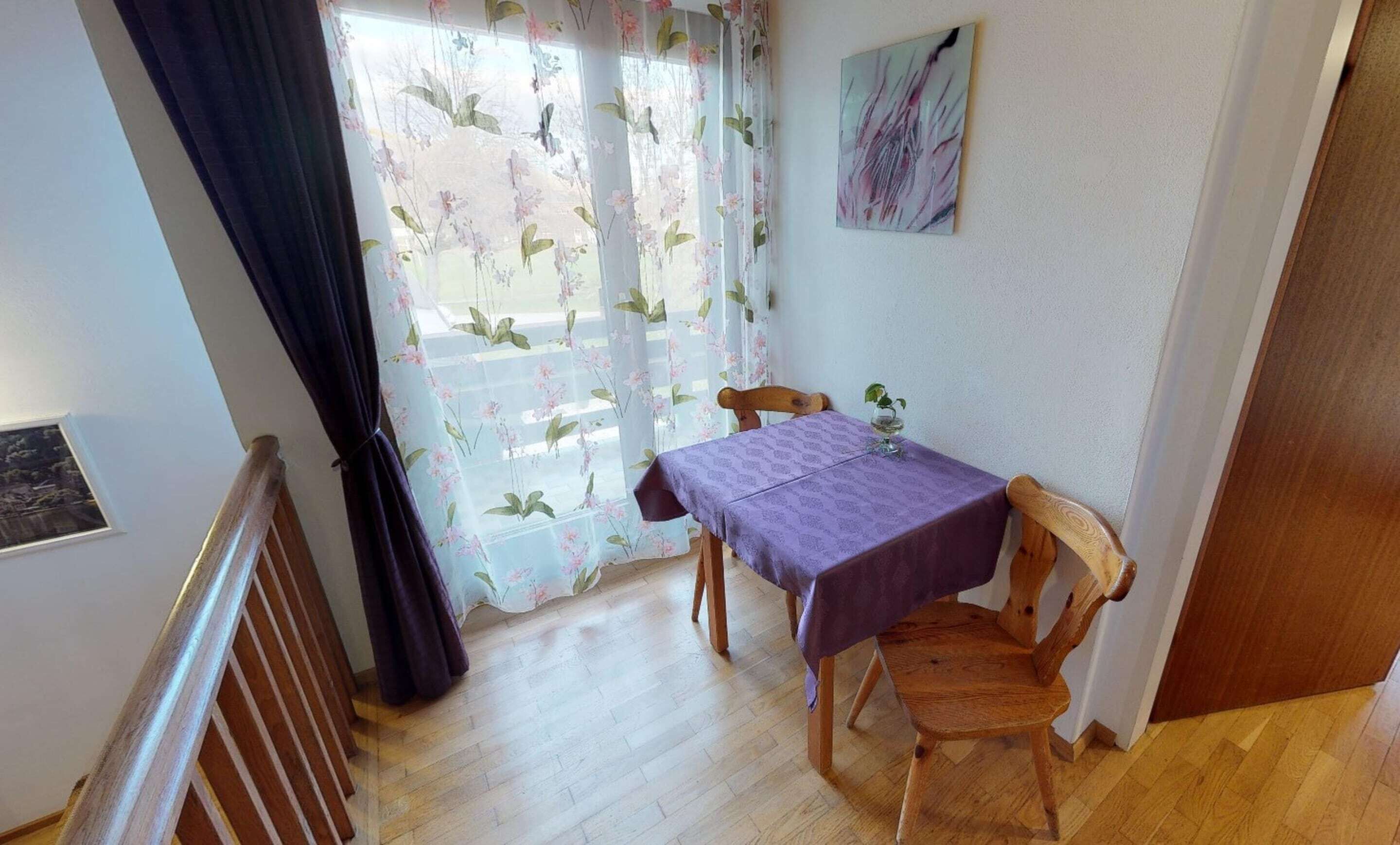 Ein kleiner Essbereich mit Tisch und Stühlen, ein Fenster mit floralem Vorhang und ein Bild an der Wand.