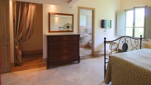 A bedroom with a wooden dresser, a bed with a floral bedspread, a mirror, a television, and a bathroom visible through an open door. - Belvilla by OYO Beautiful Farmhouse With Pool (San Gimignano)