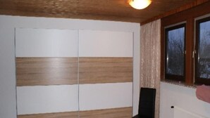 Ein Zimmer mit Holzboden, einem schwarzen Stuhl, einer Schiebetür mit Holzeinlagen und einem Fenster mit Vorhängen, sowie einem Heizkörper.