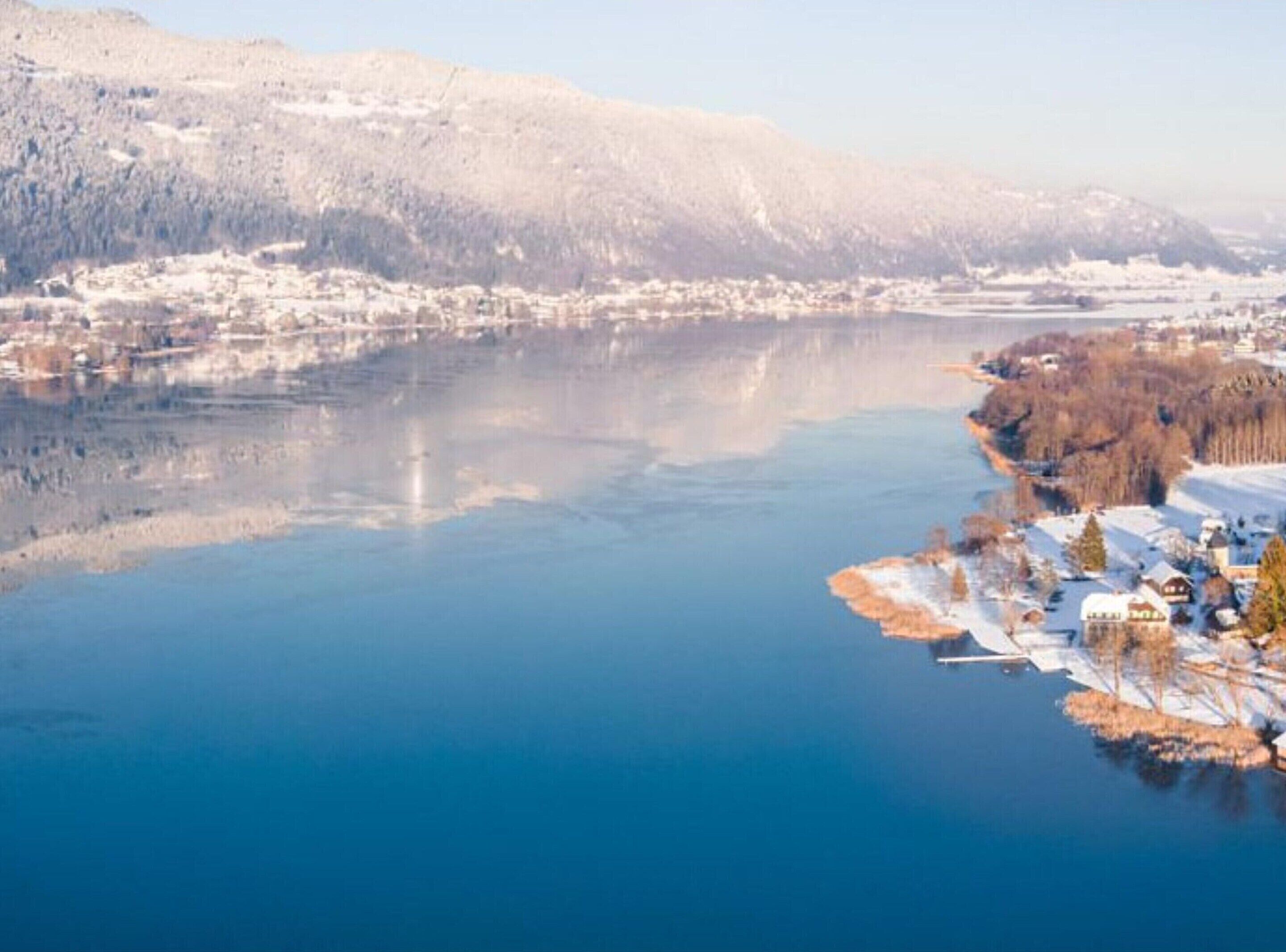 A panoramic view of a lake with snow-covered mountains in the background and a small town with houses along the shore.