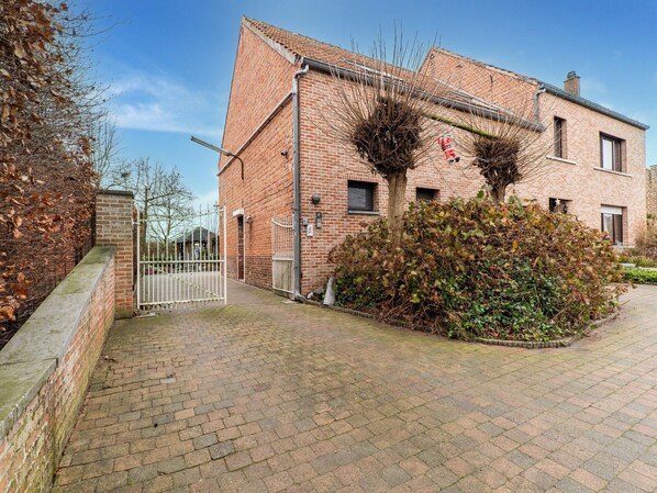 A brick building with a gated entrance, a paved driveway, and a small garden area with bushes and a tree. - Zoutleeuw Home With Pool (Zoutleeuw)