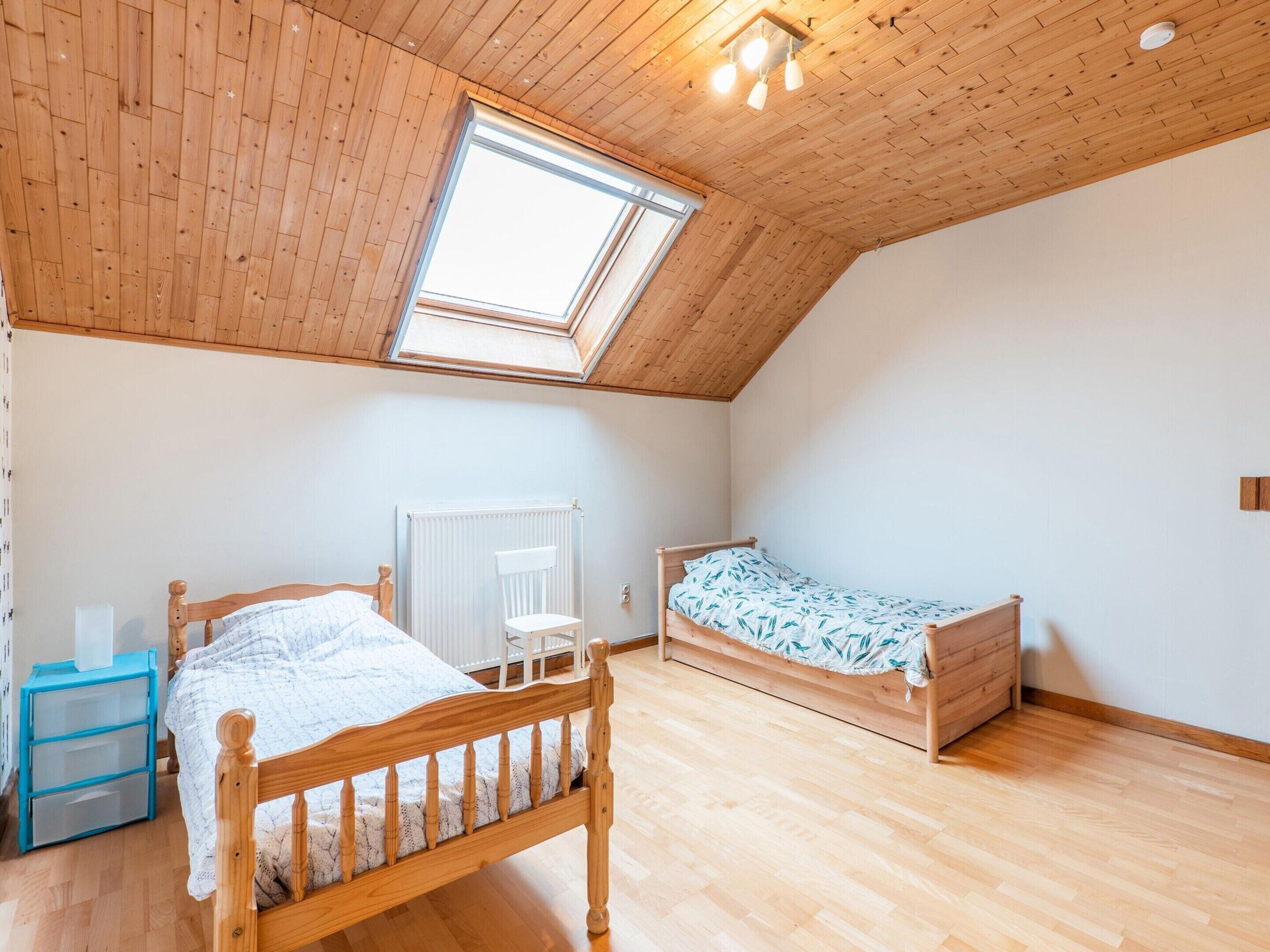 Une chambre avec un plafond en pente, un plancher en bois, un lit recouvert d’une courtepointe blanche, une chaise, un radiateur et un puits de lumière.
