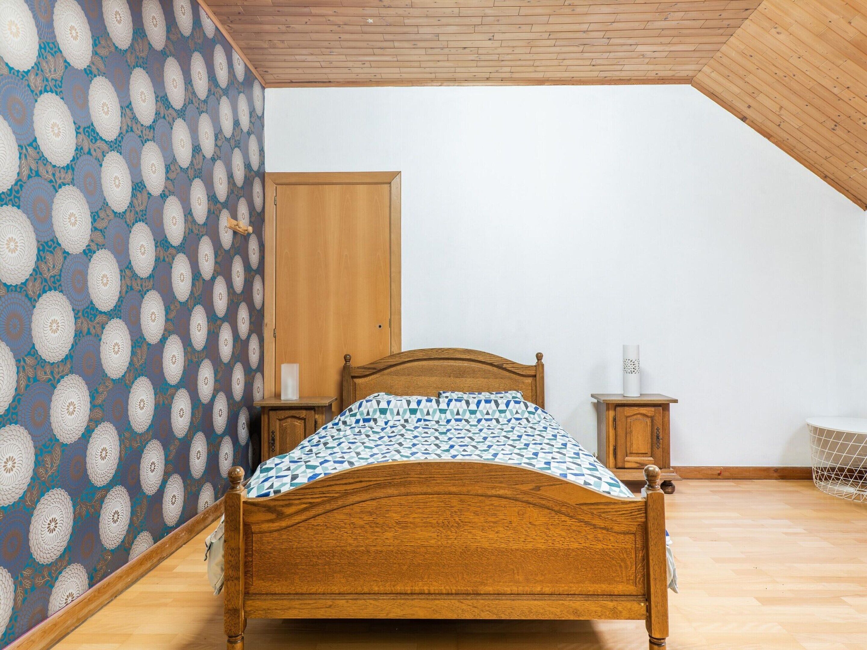 A bedroom with a wooden bed, a nightstand, a wooden wardrobe, and a patterned wallpaper.