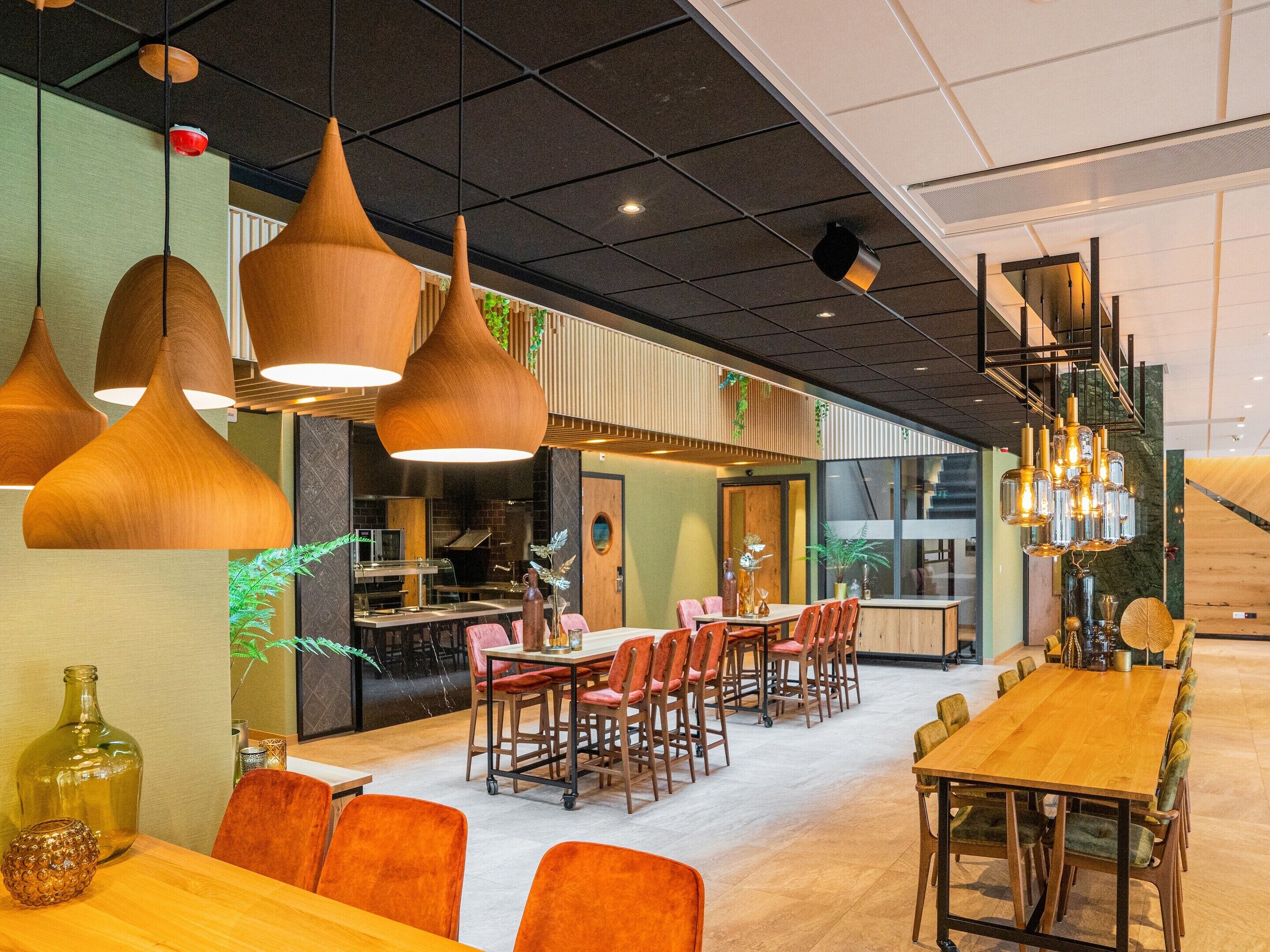 Un interior de restaurante moderno con mesas y sillas de madera, iluminación colgante y una zona de cocina a la vista.