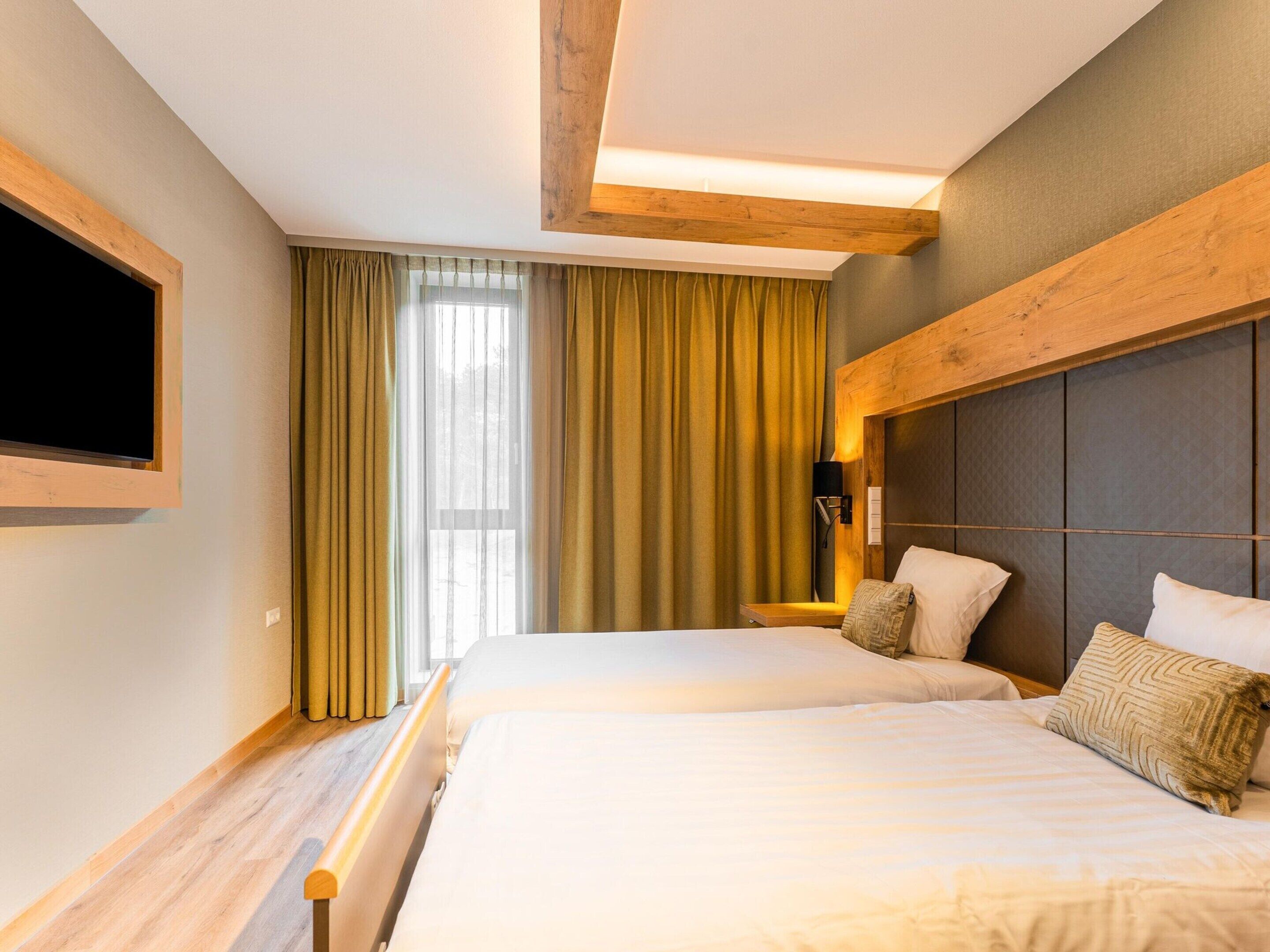 A hotel room with two beds, a large window with curtains, a mounted TV, and a wooden headboard.